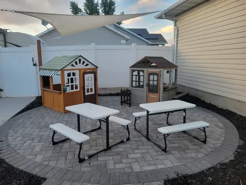 A paved patio with two kids’ picnic tables and two wooden playhouses under a shade sail