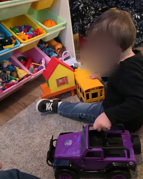 A toddler, seen from the side, playing with a toy truck and a purple toy car next to bins of toys